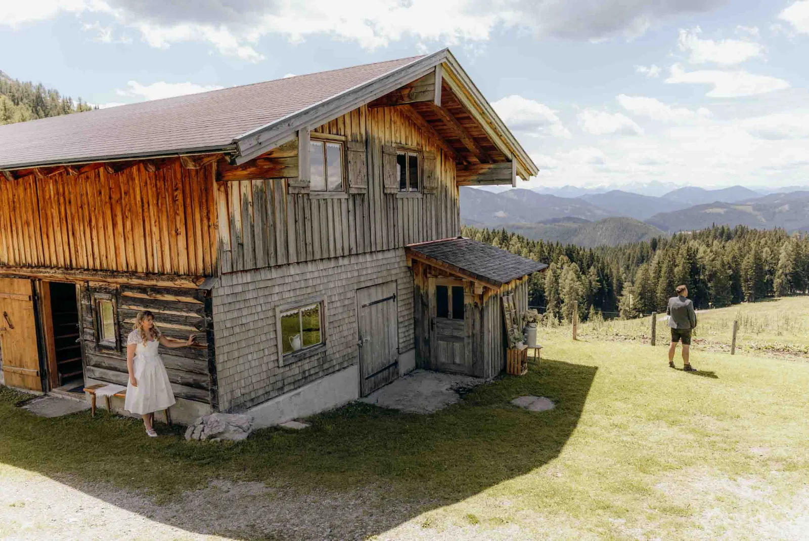 Die Braut steht entspannt an der Holzwand der Loekenwaldhuette, waehrend der Bruetigam ein Stueck entfernt wartet.