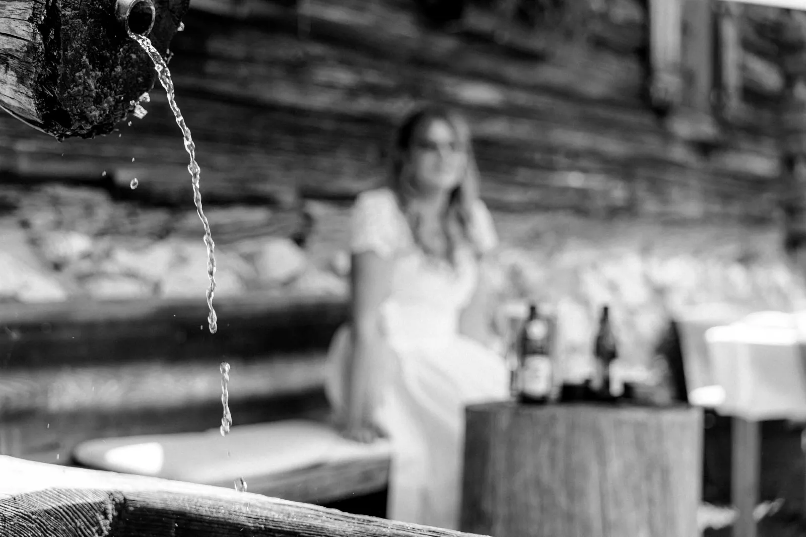 Frisches Wasser plätschert aus einem Holzbrunnen, während im Hintergrund eine Braut auf der Holzbank sitzt.