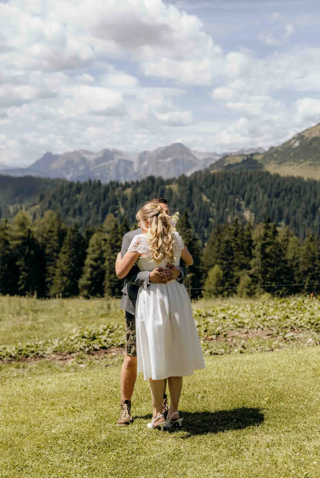 Ein Brautpaar umarmt sich herzlich auf einer Bergwiese mit weitem Blick auf die Alpen im Hintergrund.