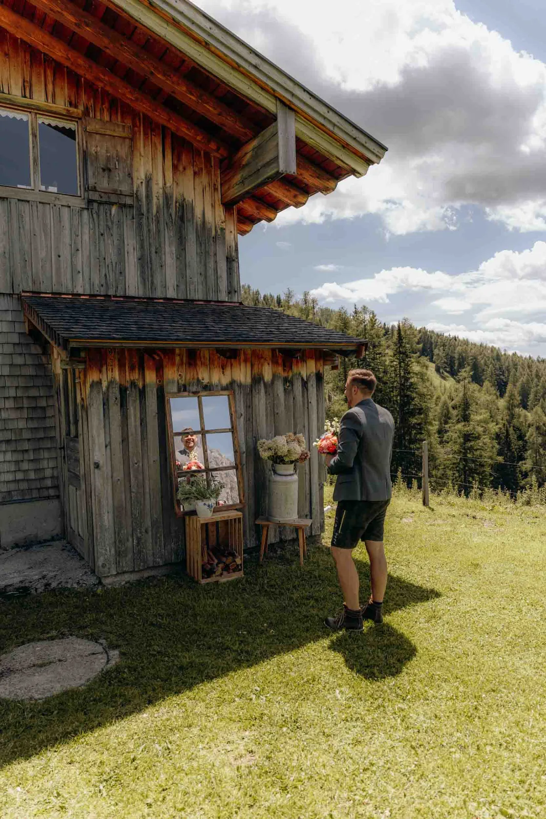 Ein Bräutigam steht mit Blumen in der Hand vor einem Spiegel an einer Holzhütte in den Bergen.