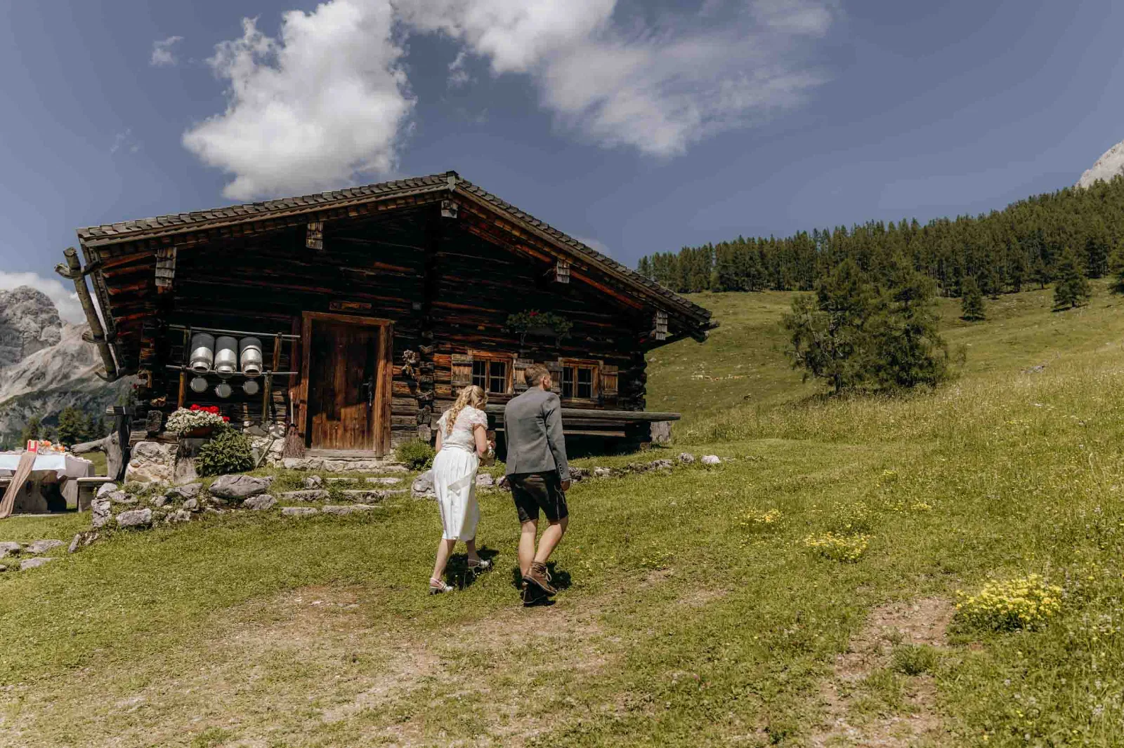 Ein Brautpaar geht gemeinsam über eine grüne Almwiese in Richtung der rustikalen Holzhütte in den Bergen.