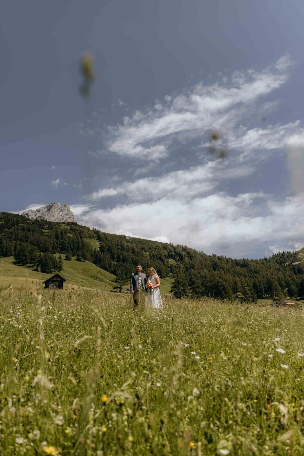 Ein Brautpaar steht auf einer weiten Almwiese mit Blick auf die Alpen, während wir diesen Moment bei der Hochzeit festhalten.